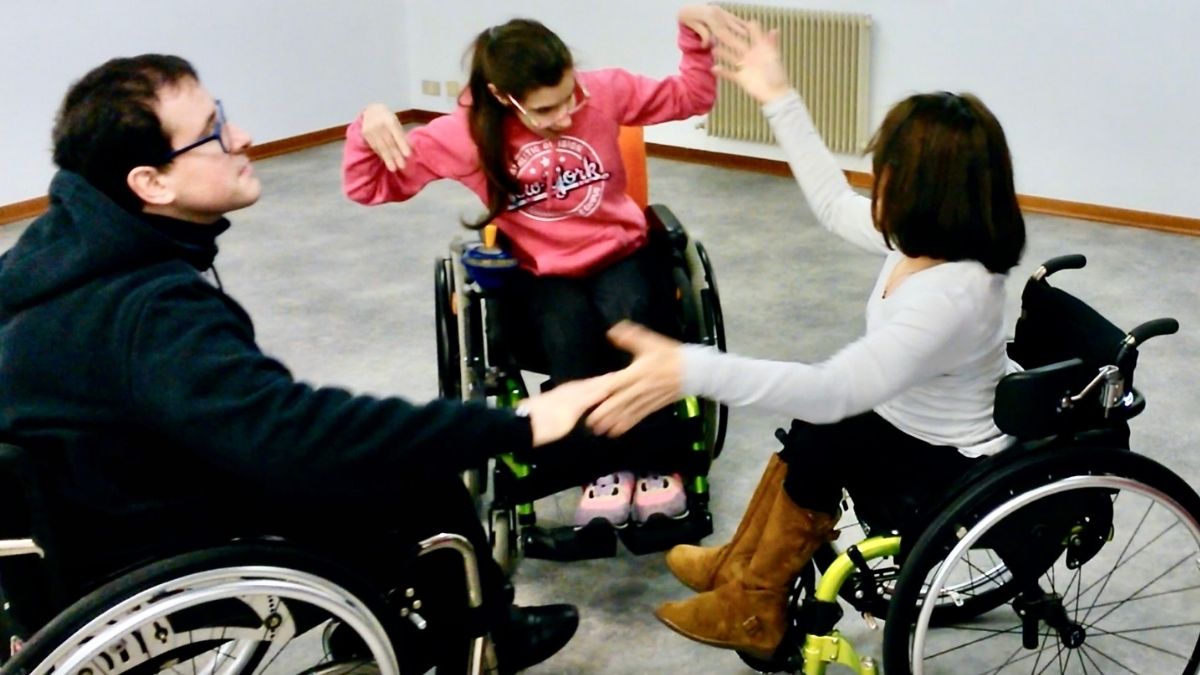 Nell'immagine ci sono tre ragazzi che sono seduti in carrozzina e stanno facendo un movimento con le braccia, esercizio di danza inclusiva. 