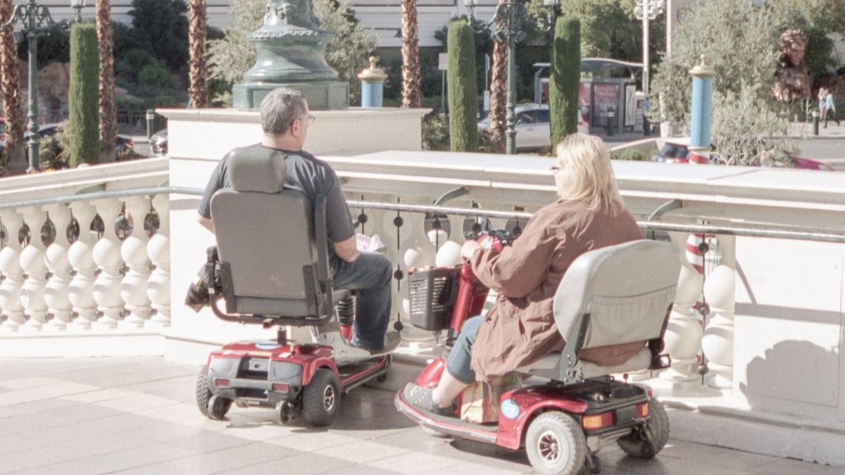 Nella foto sono presenti due persone in sedia motorizzata che si muovono in città. 