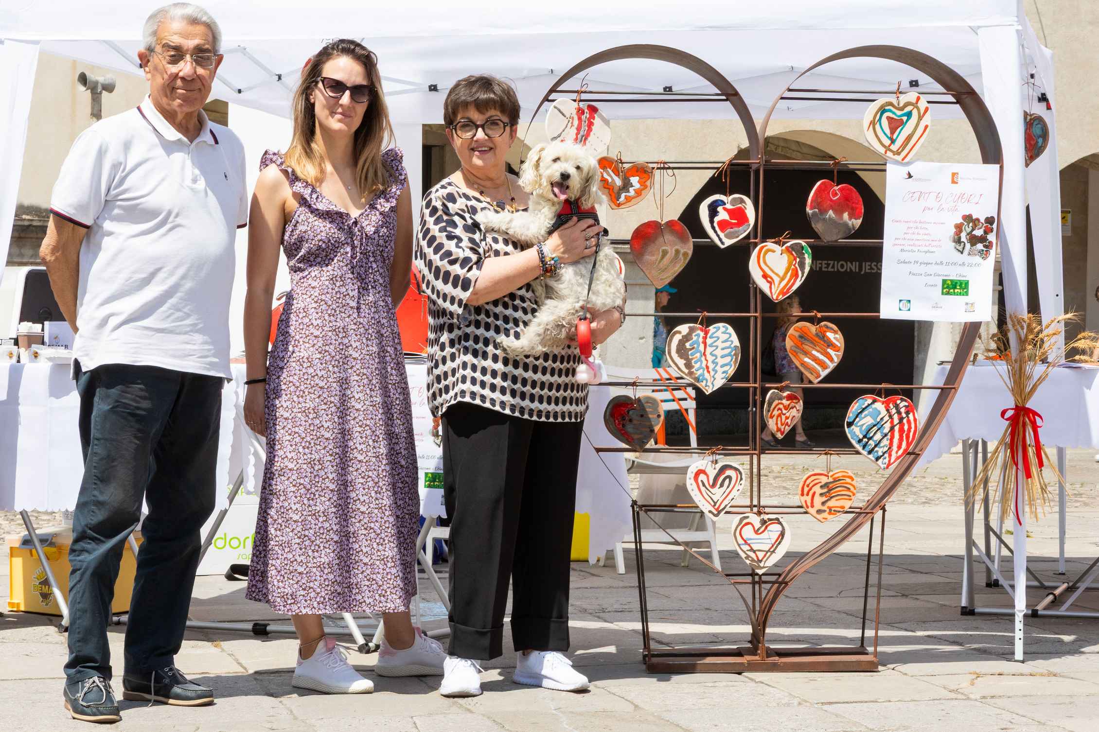 Tre persone posano davanti a uno stand all’aperto che promuove accessibilità e attenzione alle fragilità. Accanto a loro una struttura a forma di cuore decorata con cuori colorati. Una donna tiene in braccio un piccolo cane bianco.