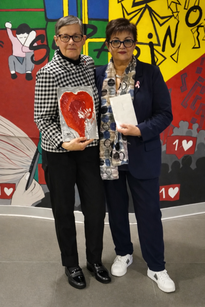 Due donne posano una accanto all’altra in uno spazio interno, davanti a una parete colorata con illustrazioni stilizzate. Una donna tiene in mano un quadro con un cuore rosso dipinto, l’altra un foglio. La scena richiama il tema delle fragilità e dell’attenzione alle persone. Entrambe guardano verso l’obiettivo.