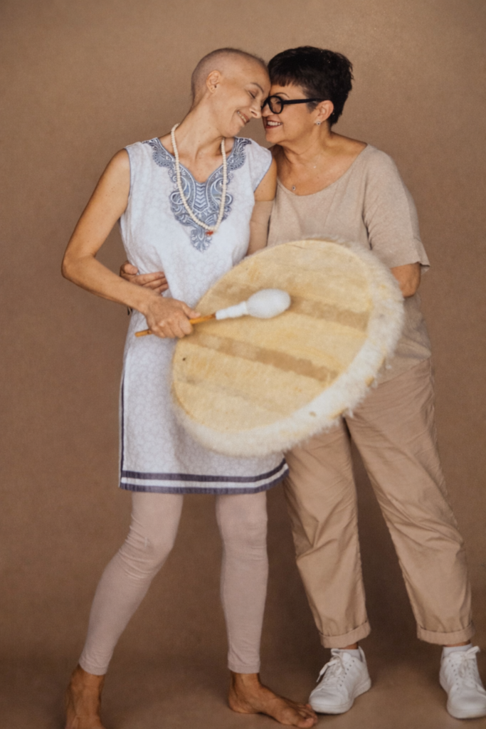 Due donne si abbracciano sorridendo, con le teste appoggiate una all’altra. Una donna tiene in mano un grande tamburo a cornice con una mazza. La scena, intima e serena, richiama il tema delle fragilità e della condivisione, su sfondo neutro.