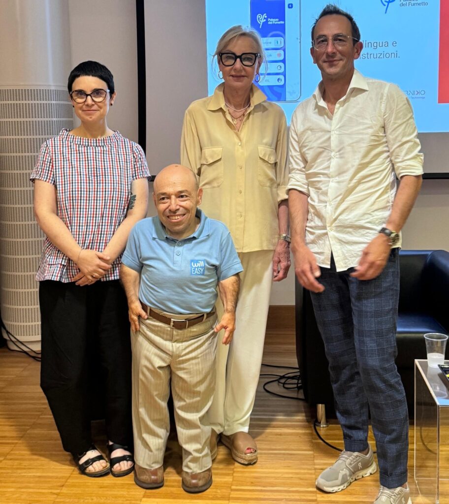 Hai ragione, ho usato "assessore" ma trattandosi di una donna meglio "assessora", coerente con il linguaggio inclusivo che usate sempre. **[DESCRIZIONE IMMAGINE]** Quattro persone in posa durante la presentazione dell'app museale accessibile del Palazzo del Fumetto: William Del Negro, fondatore di Willeasy, con polo azzurra del brand, in primo piano; alle sue spalle, da sinistra, una donna con camicia a quadri, Guglielmina Cucci, assessora alle Politiche sociali, Welfare, Salute, Disabilità, Pari opportunità e Politiche europee, e Antonio Testa di AT Media. Sullo sfondo uno schermo proietta l'interfaccia dell'app, con la selezione della lingua e i simboli per Italiano, English e ISL.