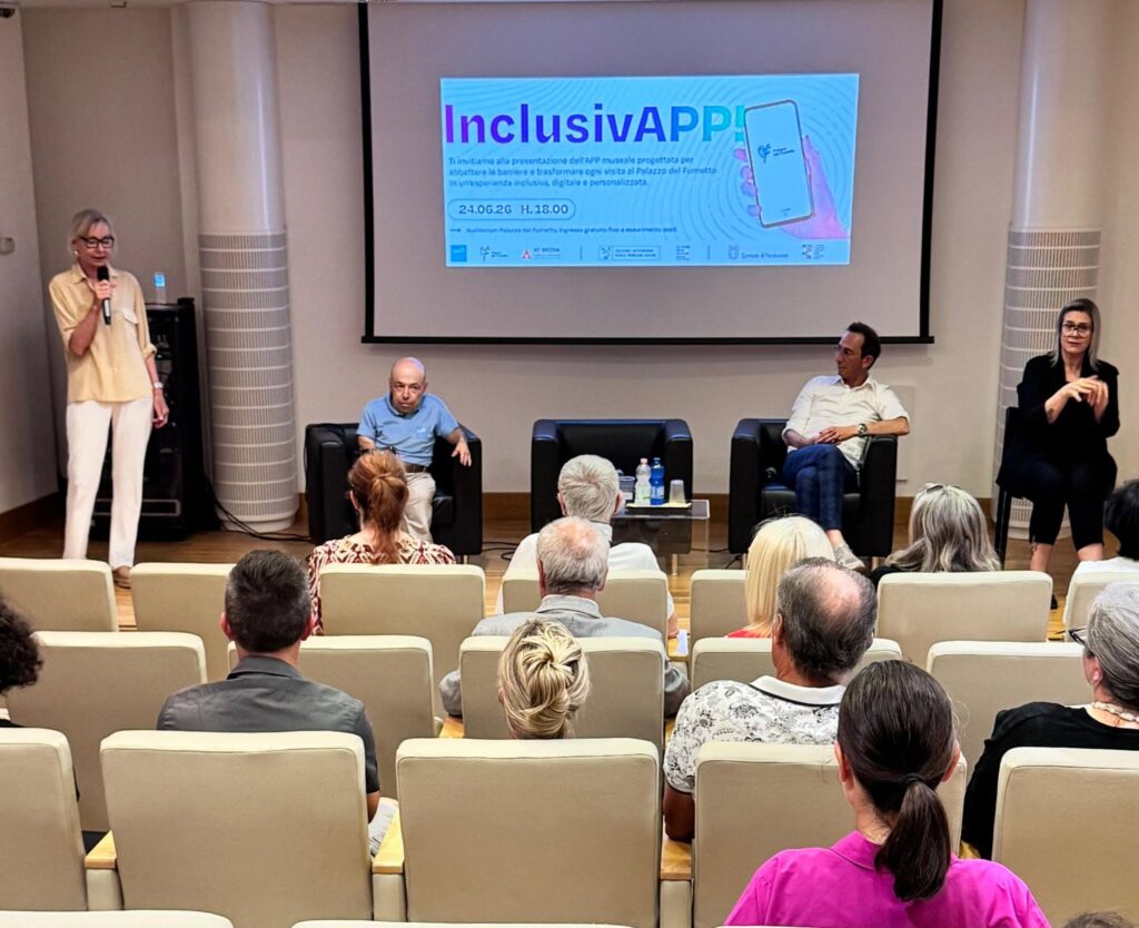 **[DESCRIZIONE IMMAGINE]** Sala auditorium del Palazzo del Fumetto di Pordenone durante la presentazione dell'app museale accessibile InclusivAPP, il 24 giugno 2026. Sul palco, da sinistra: Guglielmina Cucci, assessora del Comune di Pordenone, al microfono; William Del Negro, fondatore di Willeasy, seduto in poltrona con polo azzurra del brand; Antonio Testa di AT Media, seduto accanto a lui; a destra una interprete LIS. Sullo sfondo lo schermo con la locandina dell'evento. In primo piano il pubblico in sala, di spalle.
