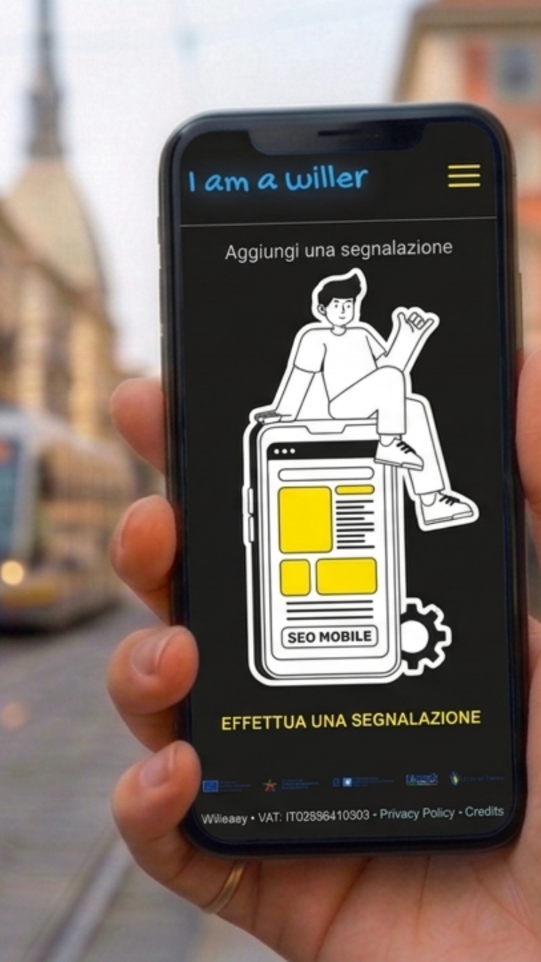 Una mano tiene uno smartphone con la schermata dell'app "I am a Willer" aperta, su sfondo nero. In alto il logo "I am a Willer" e un'icona menu a hamburger gialla. Sotto, il titolo "Aggiungi una segnalazione" e un'illustrazione di un ragazzo seduto sopra un disegno stilizzato di smartphone con blocchi di contenuto gialli e la scritta "SEO MOBILE". In basso il pulsante "EFFETTUA UNA SEGNALAZIONE" in giallo, seguito dai loghi dei partner istituzionali e, in piccolo, "Willeasy, P.IVA IT02896410303, Privacy Policy, Credits". Sullo sfondo, sfocata, una via di Torino con un tram e la Mole Antonelliana.
